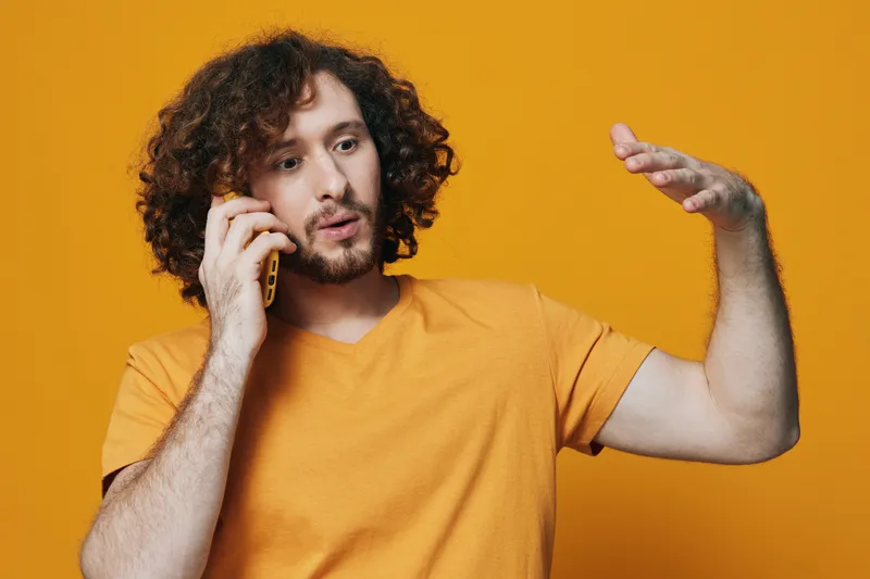 Young man with curly hair talking on the phone against a vibrant orange background, expressing excitement and engagement, wearing a casual yellow t shirt