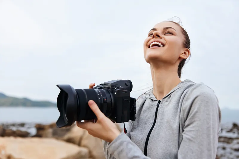 Smiling young woman enjoying photography on the beach, wearing a gray hoodie, showcasing happiness, a coastal background, ideal for summer themes