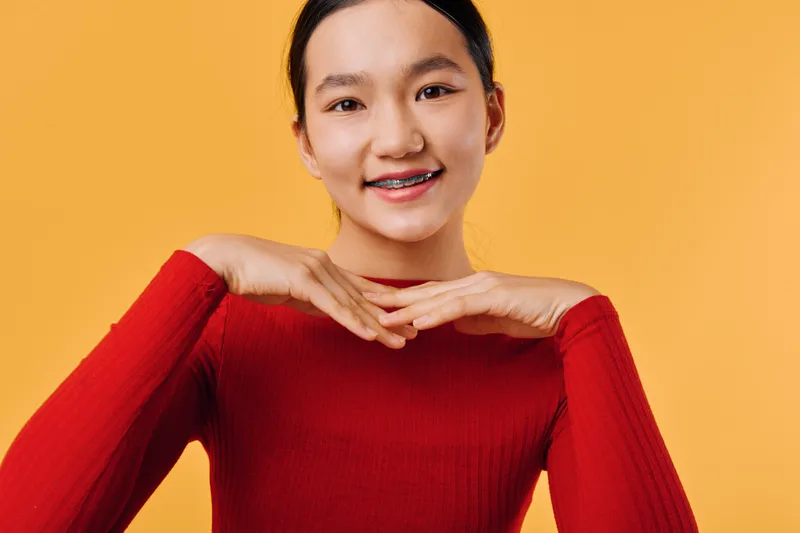 Young woman with braces smiling and posing with hands under chin against orange background. Asian female model in red sweater looking cheerful and confident with natural makeup.