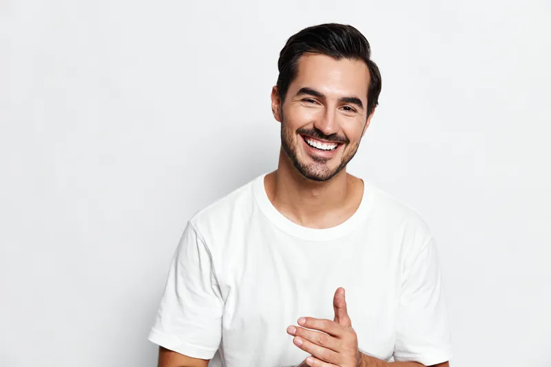 Full body smiling happy cheerful young man wearing white t-shirt, looking at the camera with a friendly expression, isolated on plain solid light background studio portrait.