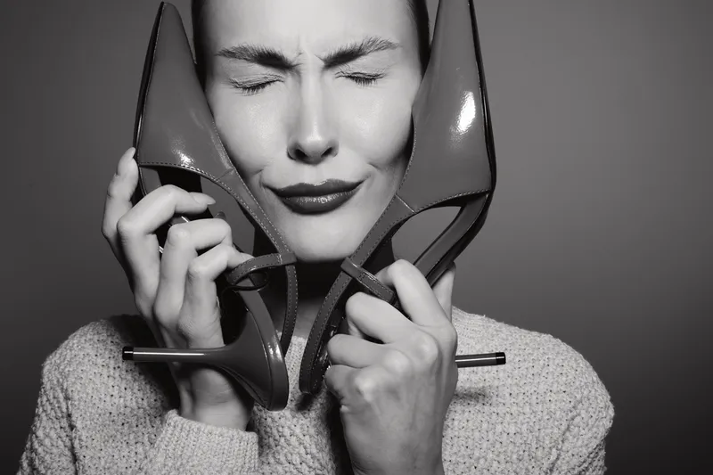 Young white woman with fair skin holds high heel shoes close to face, eyes closed with emotional expression in black and white studio portrait.