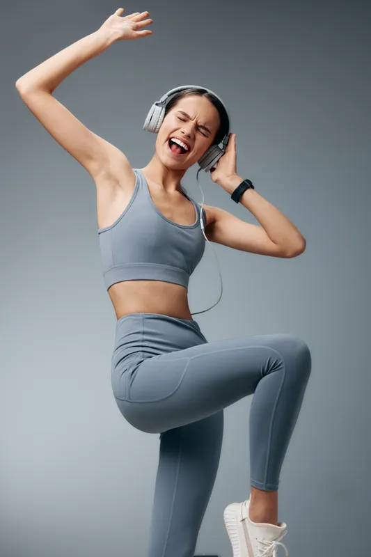 Young woman in gray fitness outfit dancing and exercising with over-ear headphones, lifting knee and arm in an energetic pose against a seamless studio gray background, joyful sports concept.