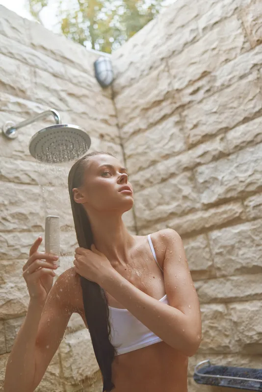 young woman showering outdoors under rain showerhead with wet hair and serene expression, natural stone wall background, wellness and relaxation concept