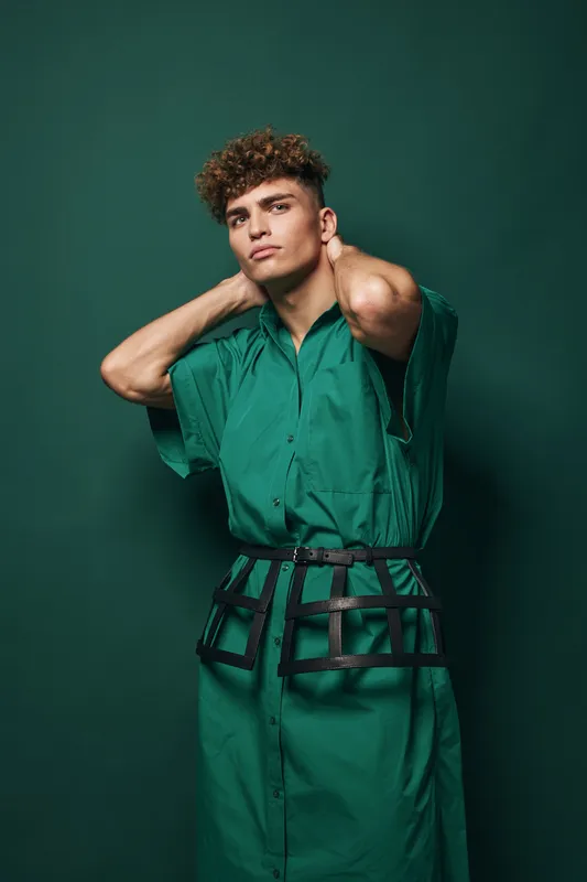 Young man with curly hair wearing green dress and black belt posing with hands behind neck against green background in studio. Fashion, style and confident model portrait.