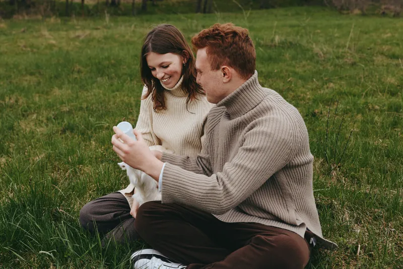 Young couple in cozy sweaters sits on grass in a meadow enjoying time together while holding a small white baby goat with smiles and relaxed mood on a cloudy day.