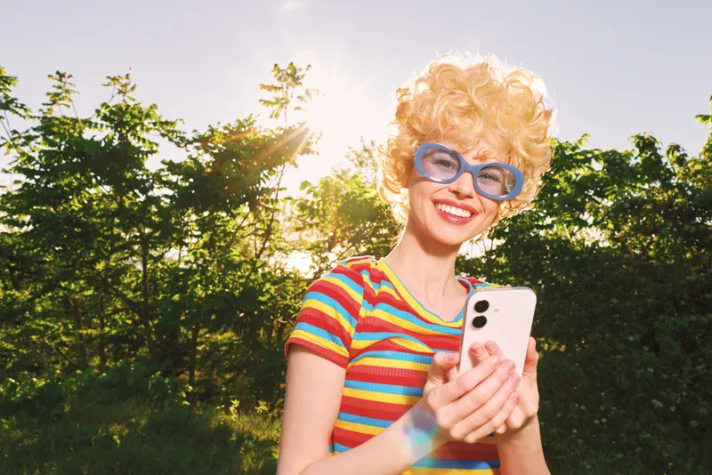 Bright outdoor portrait of a smiling woman wearing a retro striped shirt and blue glasses, taking a selfie in a sunny park, emphasizing cheerful lifestyle and retro film color
