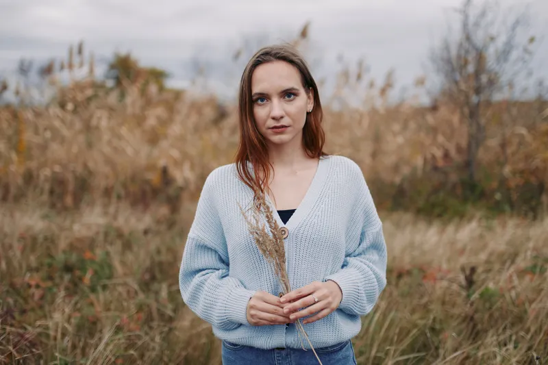 A candid portrait of a young woman standing in a grassy meadow holding dried stalks, conveying authenticity and quiet credibility through natural expression, soft light, and unposed presence.