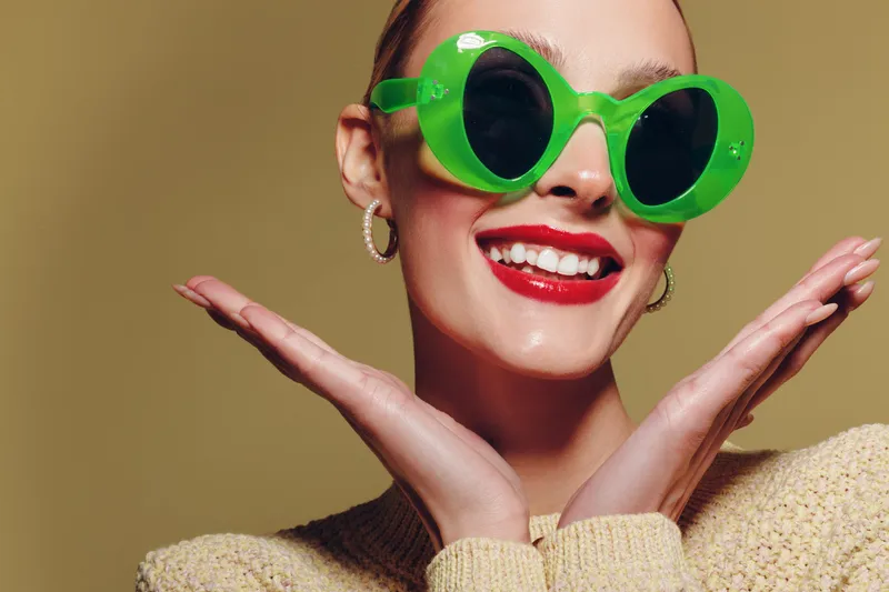 Smiling woman with bright red lipstick wearing large green sunglasses and hoop earrings poses with hands near face. Fashion portrait with beige background and joyful expression. Beauty and style