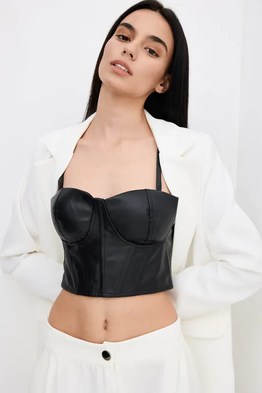 Stylish woman wearing black leather bustier and white suit jacket stands confidently against plain white background, showcasing modern fashion and minimalistic style.