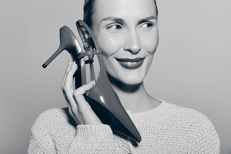 Smiling caucasian woman with fair skin holds high heel shoe near ear, wearing knitted sweater. Concept for fashion, style, and playful mood in black and white.