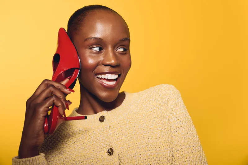 Happy dark skinned African American woman in beige sweater holding red high heel shoe near ear smiling on yellow background. Concept of fashion, joy, and playful communication.