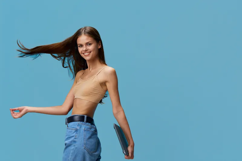 smiling woman with long hair wearing beige crop top and blue jeans holding tablet against blue background, casual style, youth lifestyle, technology accessory