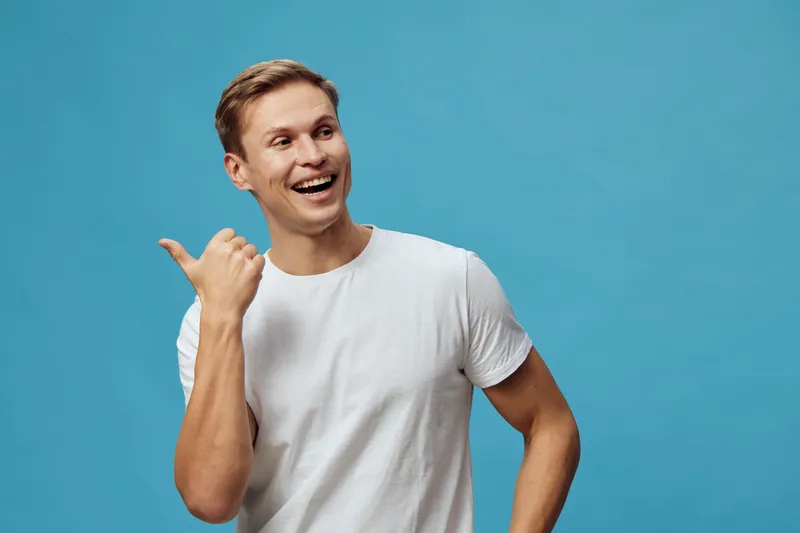 Smiling young man wearing casual white t-shirt cheerful expressing happiness hold thumb up gesture isolated on blue background studio portrait.