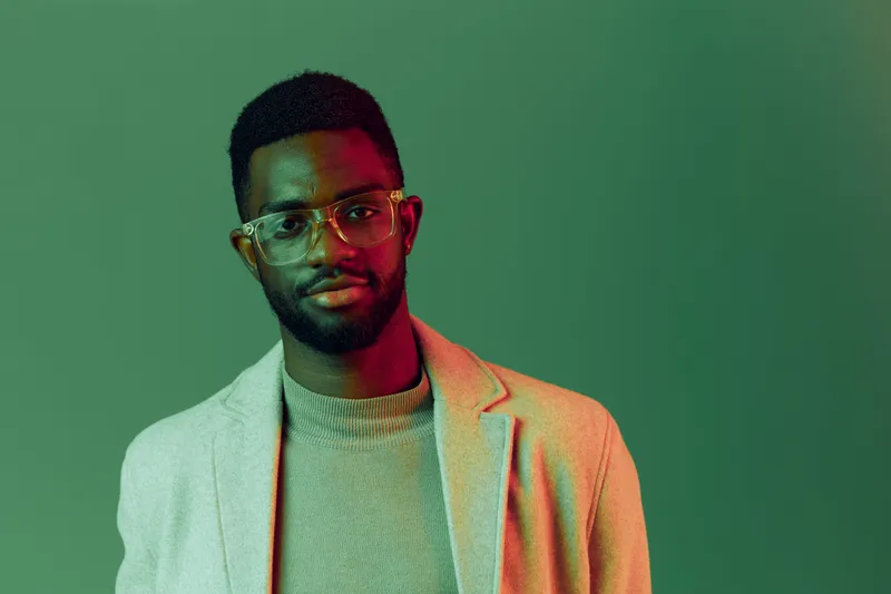 Stylish young Black man in a light gray blazer and glasses, posing against a vibrant green background, exuding confidence and modern fashion