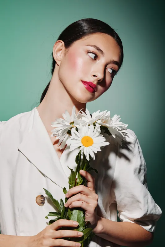 Elegant woman with smooth black hair holds white daisies while wearing a white blouse on a green background. Glow skin and red lips create a fresh beauty concept.