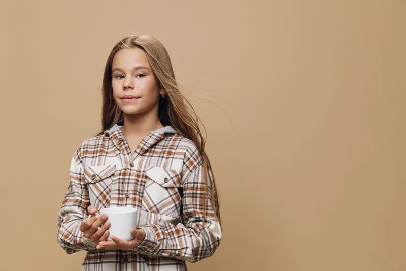 Girl with long hair holding white cup wearing checkered shirt standing on beige background indoors. Portrait of child, casual clothing and relaxed mood in studio photo.