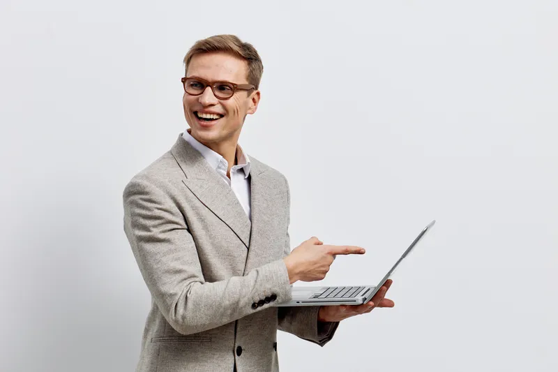 Confident smiling young man in business casual attire holding a laptop, pointing at the screen, and presenting on plain light gray background. Modern corporate image, professional, success, communicat