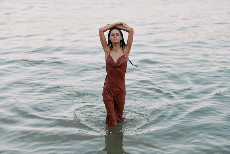 A graceful woman stands waist deep in calm water, wearing a rust colored jumpsuit. Her arms reach overhead while gentle waves ripple, creating a serene coastal moment.