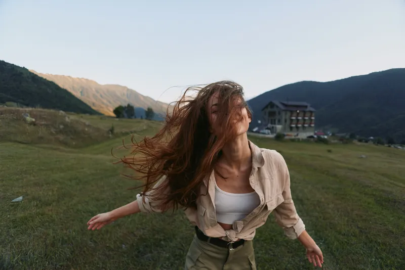 Cheerful woman outdoors in a meadow, arms open, wind in hair, sunlit scene, casual outfit, relaxed lifestyle moment, natural colors, expansive landscape