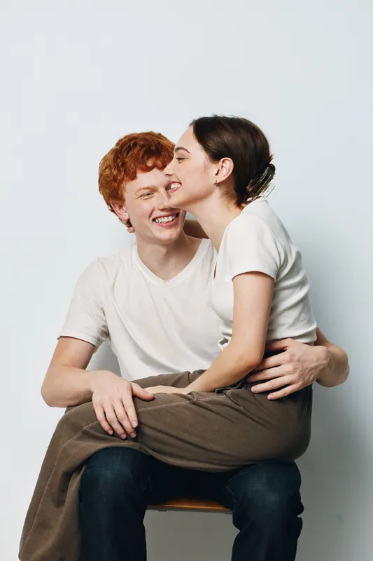 Young couple sitting together with joyful expressions, woman sitting on man's lap, happy relationship and affection in casual clothes on white background.