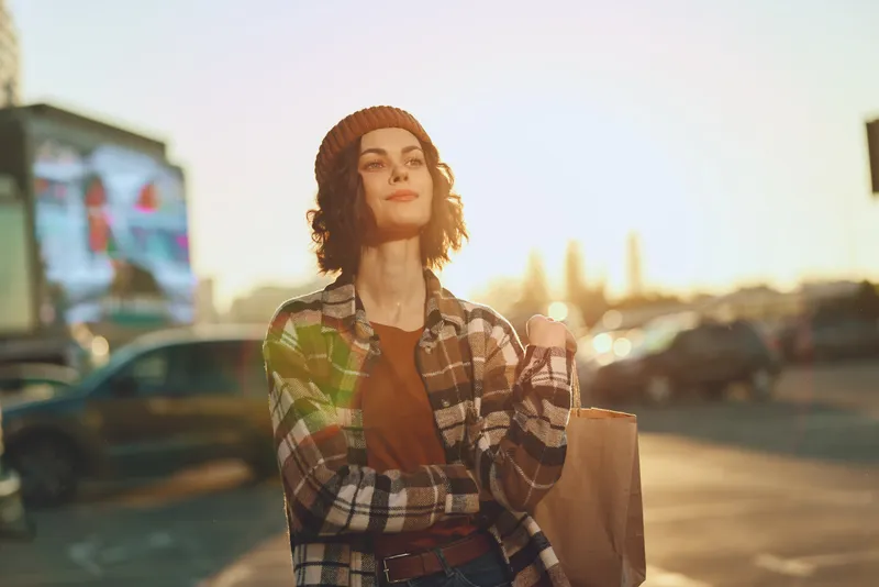 Woman shopping in urban street at golden hour, candid lifestyle portrait with authenticity and golden hour glow, holding a paper bag with confident expression and emotional storytelling vibe.