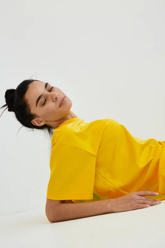 Young woman with closed eyes wearing a vibrant yellow t-shirt lying relaxed on a white floor, showcasing calmness and minimalistic indoor lifestyle mood.