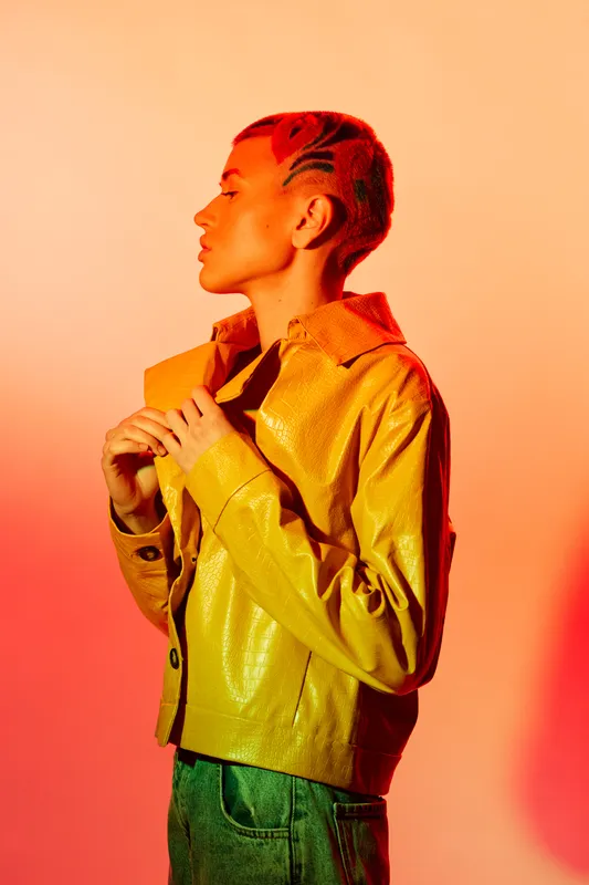 Profile of a stylish woman with braided hair wearing a yellow jacket and green pants, illuminated by warm orange lighting creating a modern, vibrant portrait.