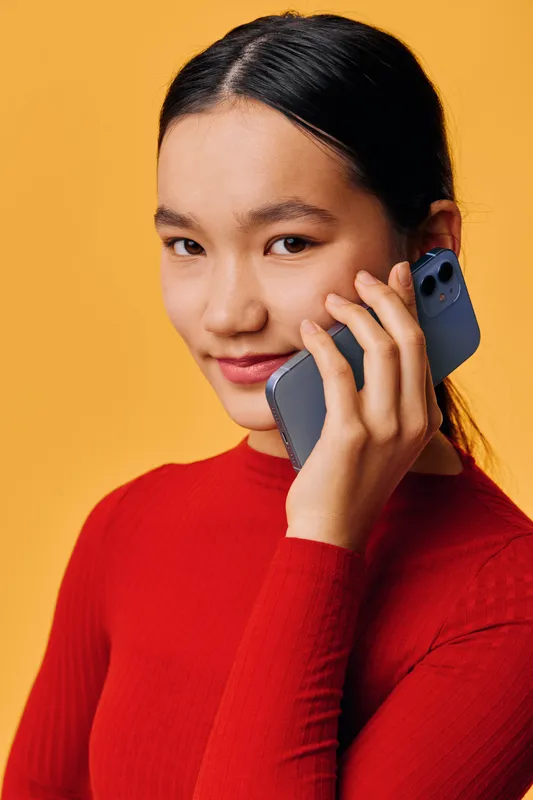 Young woman in red sweater holding smartphone close to ear while smiling and making a call. Studio portrait with warm yellow background and casual hairstyle for communication concept.