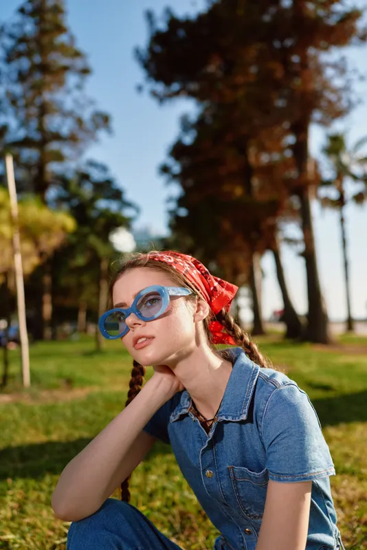 Lifestyle woman in modern boho-western denim street style wearing blue sunglasses and red bandana outdoors in natural park under warm film color tones.