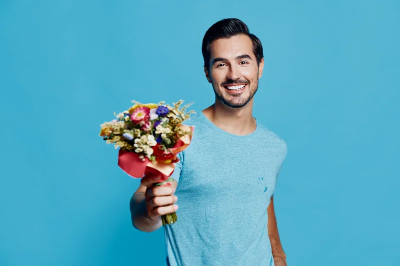 Happy smiling young man holding a bouquet of flowers, wearing a light blue t-shirt, isolated on a solid blue background in a cheerful studio portrait.