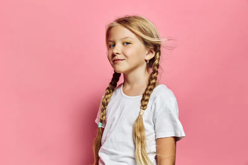Smiling young girl with long blonde braids wearing a white t-shirt poses confidently against a pink background. Studio portrait with natural light and casual style.