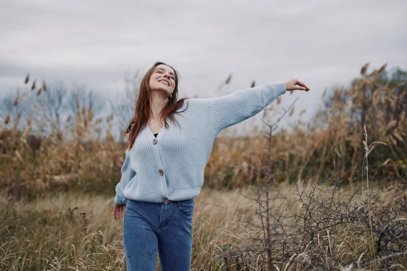 Authentic moment of a woman outdoors, wearing a knit sweater, smiling with arms outstretched, capturing genuine happiness and natural grace in an open field.