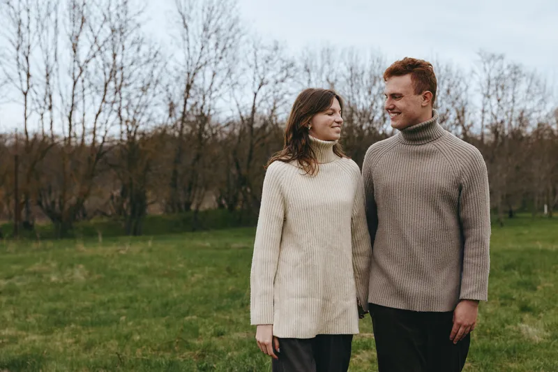 Couple outdoors strolls through a quiet park in knit sweaters, sharing warm smiles and gentle contact, enjoying a calm lifestyle amid bare trees and open fields, romantic connection