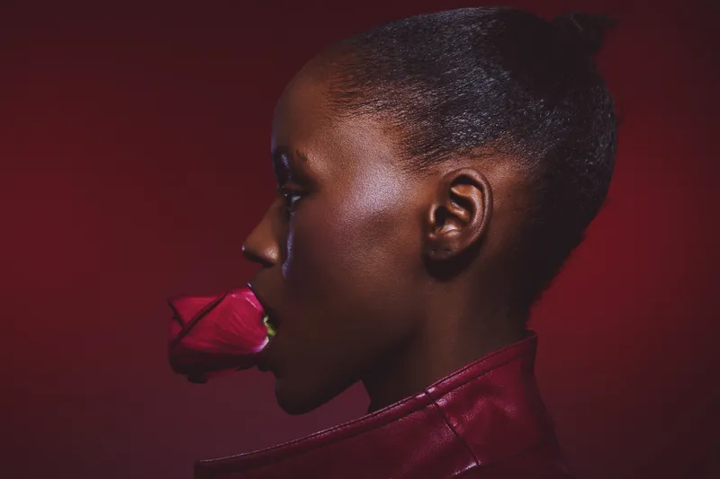 Profile of black African woman with dark skin holding red rose in mouth wearing red leather jacket. Intense dramatic lighting on deep red background. Concept of beauty, fashion, and elegance.