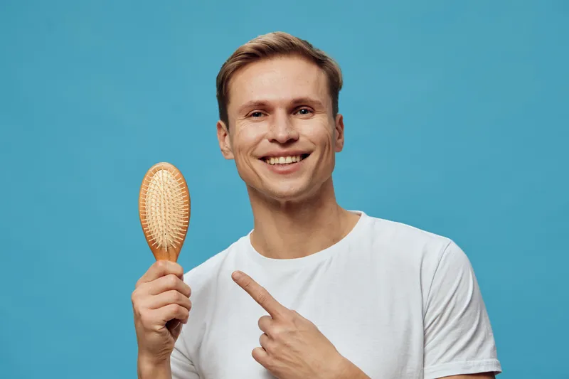Smiling man with short brown hair wearing white t-shirt holding wooden hairbrush pointing at it on blue background studio portrait. People lifestyle concept