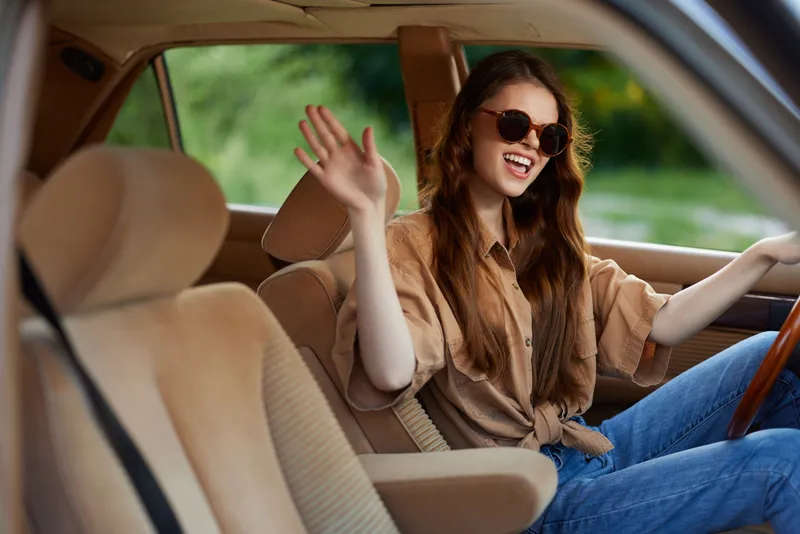Young woman with long hair waving from a car, wearing sunglasses and a stylish outfit, exuding joy and confidence in a natural green setting.