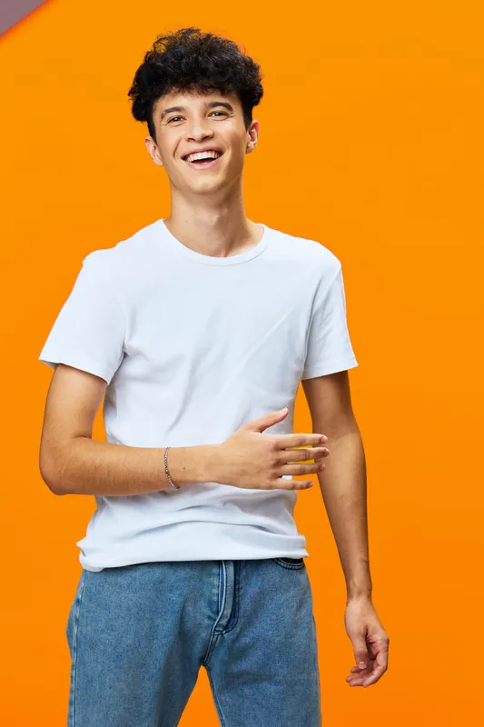 young man laughing in casual white t-shirt and jeans with curly hair, standing against bright orange background, joyful and relaxed mood
