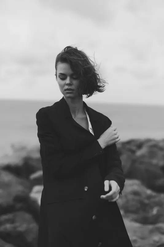 Black and white photo of a woman in a dark coat standing by rough rocks along the sea, wind in hair, moody atmosphere