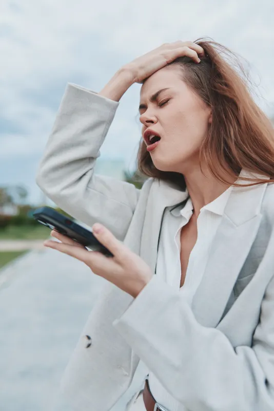 frustrated woman in light jacket holding smartphone and expressing stress outdoors with cloudy sky background