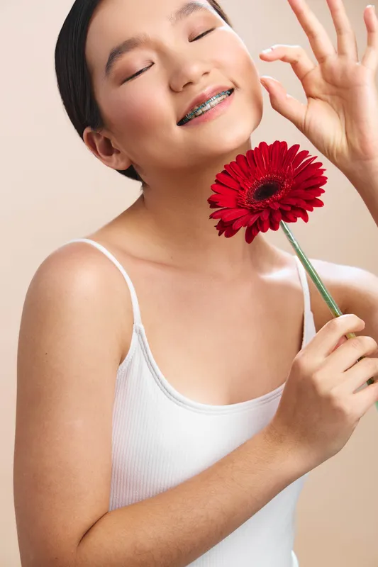 asian woman smiles softly in a bright studio, holding a red flower near her face, expressing calm joy and graceful charm through a relaxed, candid portrait.