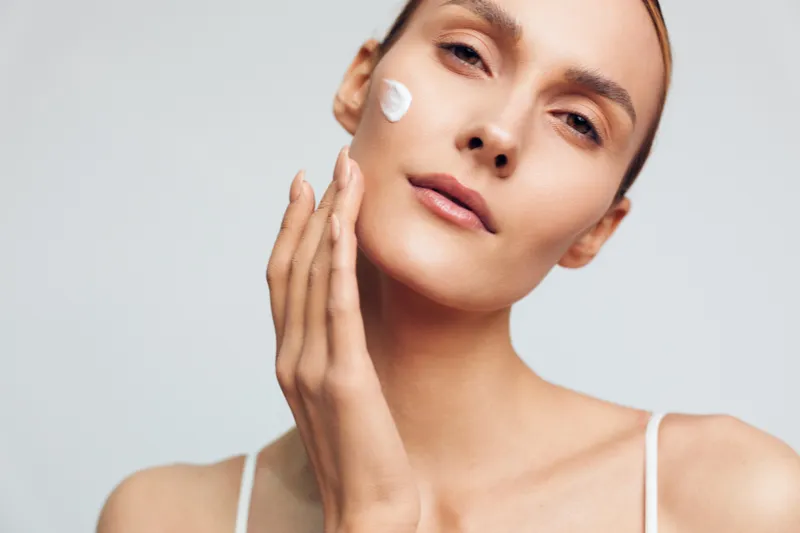 Young woman applies face cream on clean skin with gentle touch isolated on white background, natural skincare beauty routine concept for healthy glowing complexion.