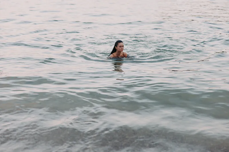 A solitary swimmer rises from calm sea water, ripples radiating outward as soft light plays on the surface, capturing a moment of quiet strength and graceful effort in open water.