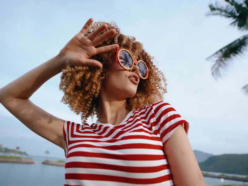 A stylish woman with curly hair wearing a red and white striped shirt and round sunglasses stands outdoors near water, shielding her eyes with a hand against a bright sunny sky.