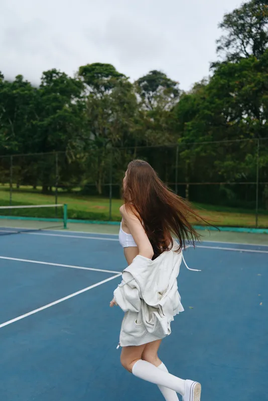 Young woman enjoying a playful moment on a tennis court, wearing sporty attire and showcasing long, flowing hair against a lush green background