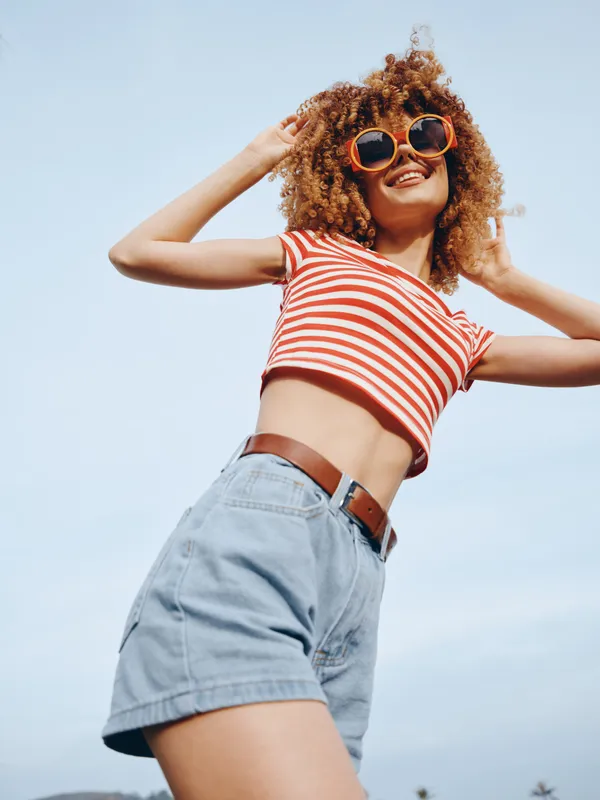 Bright and carefree portrait featuring a smiling woman with curly hair, red striped crop top and denim shorts, posed against a clear blue sky.