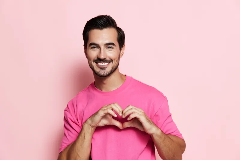 Happy smiling man wearing a pink shirt makes a heart shape with his hands against a solid pink background. Joyful, cheerful, love concept.