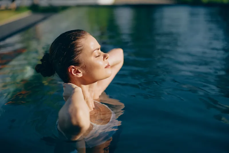 Joyful woman in a sunlit pool, eyes closed, serene pose, water ripples softly, outdoor setting, warm tones, relaxed lifestyle moment, fresh air and natural colors