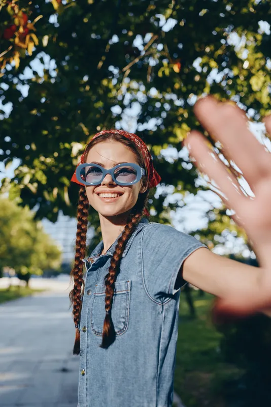smiling woman with braids wearing blue sunglasses and denim dress outdoors in sunny park with green trees during summer season