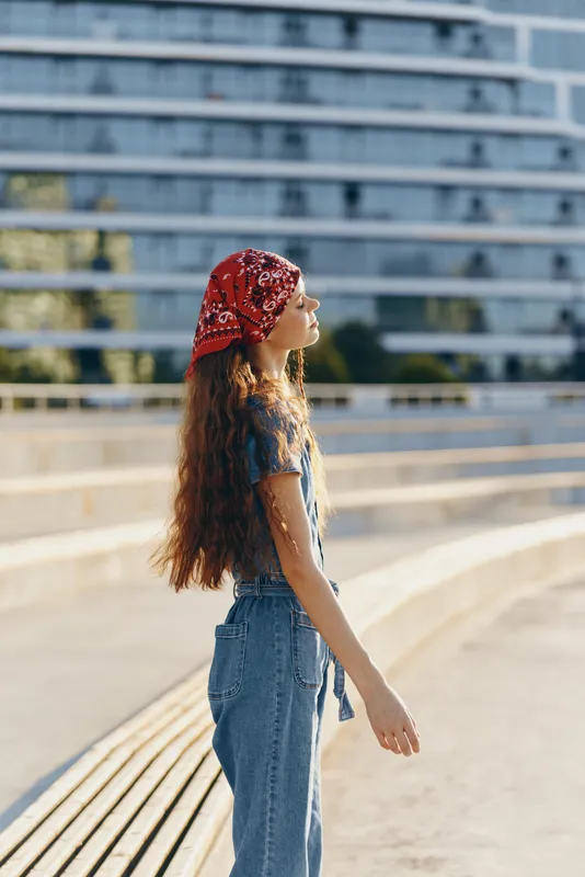 Lifestyle woman wearing modern boho-western denim street style with long hair and red bandana, captured in warm film color tones during a sunny day outdoors.