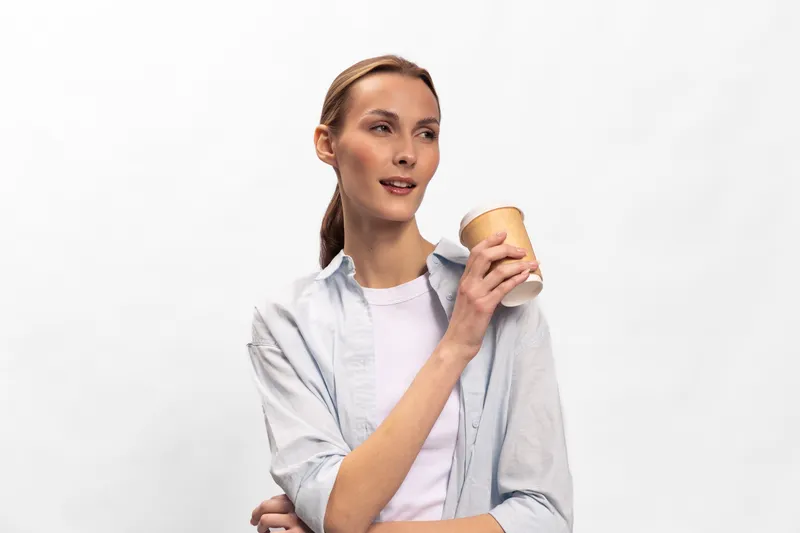 Young woman in casual shirt holding disposable cup with coffee looks aside with thoughtful expression on white background. Concept of break, beverage, relaxation, and lifestyle.